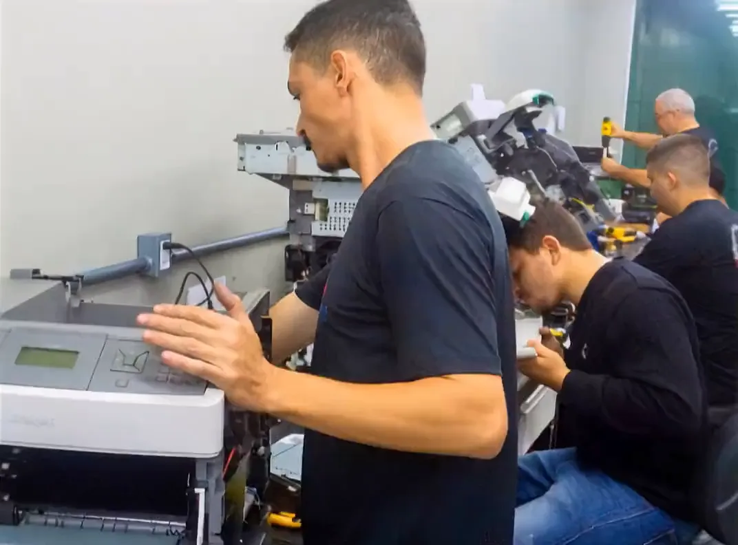 Técnico ajustando uma impressora na bancada da oficina, com outros equipamentos enfileirados ao fundo.
